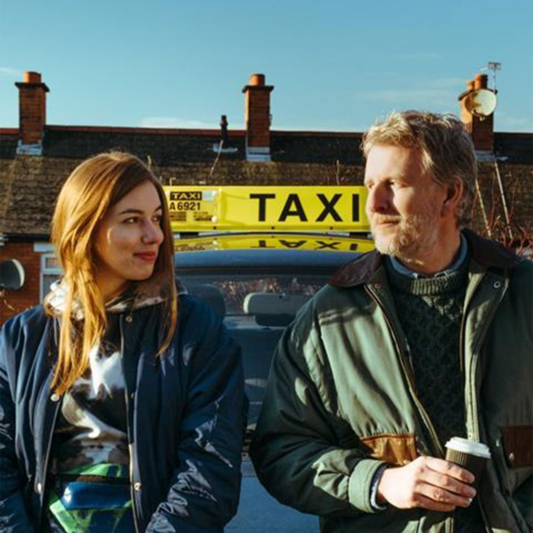 Ballywalter hero image: Eileen(Seána Kerslake) on the left, Shane(Patrick Kielty) on the right, both leaning up against a black car. A bright yellow sign saying "TAXI" is on the roof.